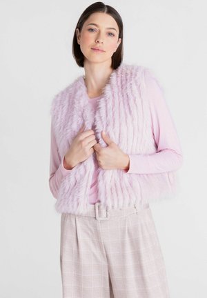 Vrouw met donker haar, die een lichtroze top met lange mouwen, een donzig roze vest en beige geruite hoge taille broek draagt en de kraag van het vest vasthoudt.