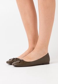 Gambe che indossano ballerine in pelle marrone scuro con piccoli fiocchi, in piedi su uno sfondo bianco semplice.