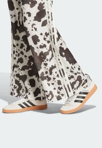 Vita byxor med ett brunt prickigt mönster och tre vita ränder på sidorna, i kombination med krämfärgade sneakers med svarta ränder och en texturerad sula.