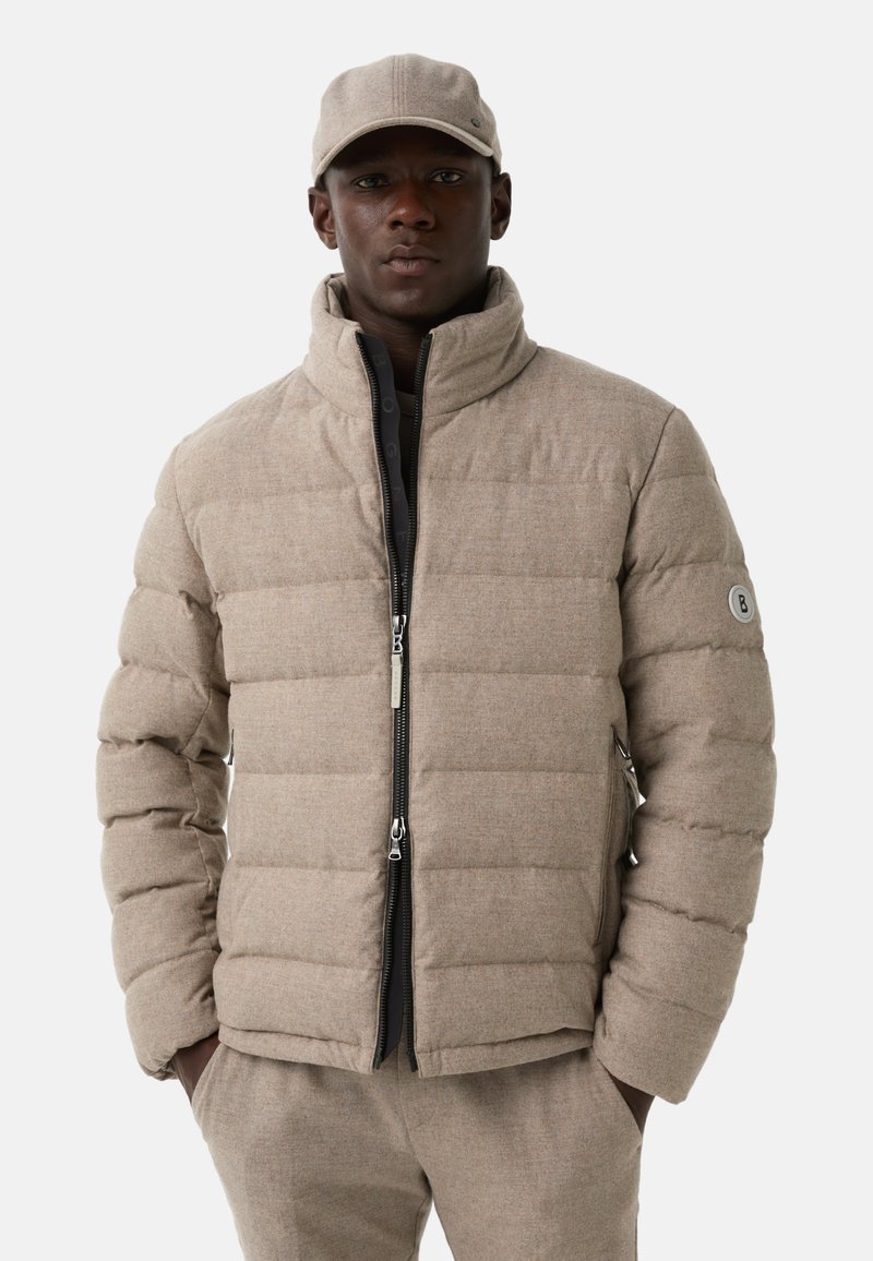 Beige pufferjack met een hoge kraag, een gestept ontwerp en een rits. Heeft zijzakken en een logo-patch op de mouw.