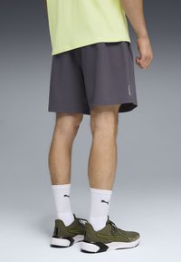 Άνδρας που φοράει σορτς σκούρου γκρι χρώματος, λευκές κάλτσες Puma μέχρι τον αστράγαλο και χακί αθλητικά παπούτσια Puma, στέκεται σε γκρι φόντο.