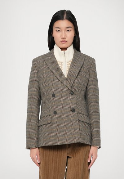 Giovane donna che indossa un blazer a quadri doppiopetto sopra un maglione a zip in maglia color crema e pantaloni in velluto a coste marroni, in piedi contro uno sfondo semplice.