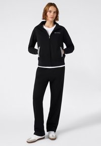 Felpa nera con zipper e colletto alto, con dettagli grigi chiari e tasche. Abbinata a pantaloni della tuta neri e scarpe da ginnastica bianche.