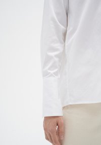Camisa blanca de manga larga con una textura suave, presenta un dobladillo con puños y un corte entallado. La tela parece ligera y ligeramente reflectante.