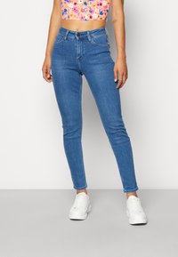 Jasnoniebieskie dopasowane dżinsy uszyte z denimu, z wysokim stanem, klasycznym pięciopocketowym designem i delikatnym wytarciem. Noszone z białymi trampkami.