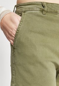 Pantalonii din bumbac de culoare verde măslin prezintă o buzunar frontal cu cusături contrastante și un tiv standard, evidențiind o textură netedă, mată.
