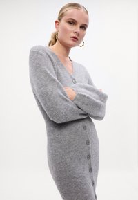 Abito grigio lavorato a maglia con scollo a V, maniche lunghe a vestibilità ampia e sei bottoni lungo il lato, che mette in mostra una consistenza morbida.