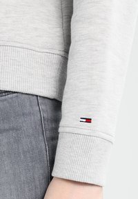Manica di felpa grigia con texture a coste e un piccolo logo di Tommy Hilfiger con strisce rosse, bianche e blu vicino al polsino.