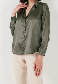 Olive green satin blouse with long sleeves, pointed collar, and button front. Designed with a smooth texture and a polished finish.