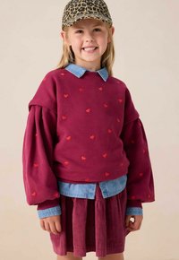 Bordeaux sweatshirt met rode hartpatronen, oversized mouwen, gelaagd over een licht spijkerhemd, gecombineerd met een bordeaux corduroy rok.