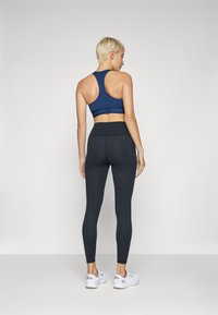 Soutien-gorge de sport bleu marine avec un dos nageur, associé à un legging noir. Les deux sont fabriqués dans un matériau extensible ; les leggings ont une taille haute, chaussures blanches.