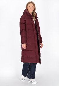 Bordeaux puffer jas met een capuchon, voorzien van een gestikt ontwerp, een voorkant met rits en zijzakken. Draagt over donkere jeans en witte sneakers.