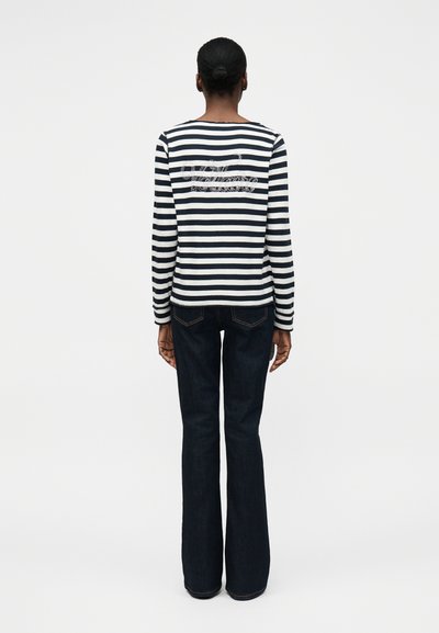 Άτομο με σκούρα μαλλιά σε κότσο που φοράει μακρυμάνικη μπλούζα με ασπρόμαυρες ρίγες και σκούρα μπλε τζιν, στραμμένο προς τα πίσω σε απλό φόντο.