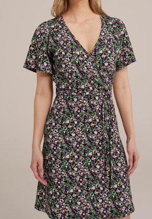 Vrouw draagt een wikkeljurk met korte mouwen met een kleine bloemenprint in groen, roze, blauw en zwart op een beige achtergrond.