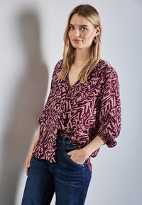 Paarse en crèmekleurige blouse met een patroon, v-hals, lange mouwen en een losse pasvorm, gecombineerd met donkerblauwe jeans. Glad materiaal, ontspannen ontwerp.