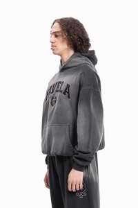 Νέος με σγουρά μαλλιά, φοράει σκούφη γκρι φούτερ και αντίστοιχα παντελόνια φόρμας με γραφικά λογότυπα, στέκεται προφίλ σε λευκό φόντο.