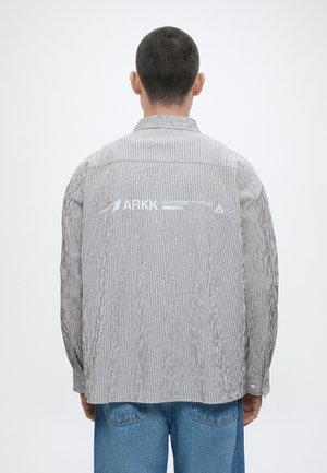 Άνδρας με γκρι και λευκό ριγέ μπουφάν με λευκό κείμενο στην πλάτη και μπλε τζιν, που κοιτάζει προς τα πίσω σε απλό φόντο.