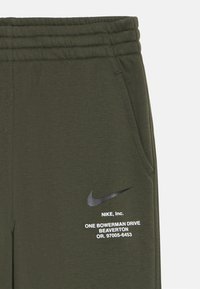 Žalios medvilnės mišinio kelnės su šoninėmis kišenėmis, elastingu juosmeniu ir juodu Nike logotipu su baltu tekstu priekyje.
