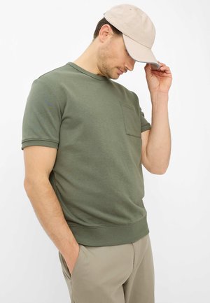 Homme portant une chemise à manches courtes vert olive avec une poche, une casquette beige et un pantalon beige, regardant vers le bas avec une main ajustant la visière de la casquette.
