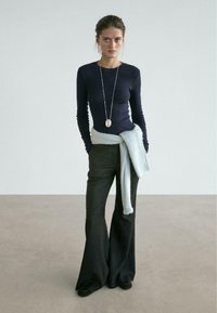 Marineblaues, langärmeliges Oberteil mit lockerem Schnitt, kombiniert mit schwarzen Schlaghosen. Hellblauer Pullover um die Taille gebunden, silberne Anhängerkette.