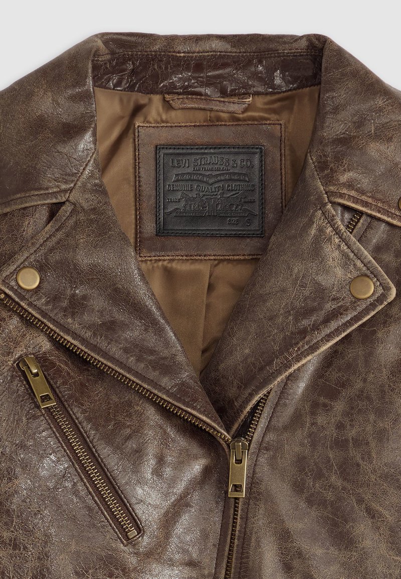 Veste de moto en cuir marron avec un col cranté, des accessoires en laiton, deux poches zippées et une surface texturée. Doublure intérieure en tan.
