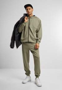 Oliwkowy, rozpinany hoodie i dresy z miękkiego materiału, w zestawieniu z czarną kurtką puchową i białymi sneakersami.