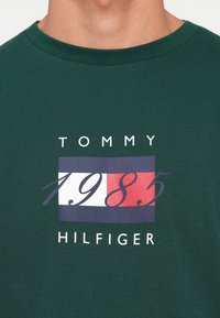 Grøn bomulds T-shirt med et grafisk design, der viser teksten "TOMMY" og "HILFIGER" samt året "1985" i rød og blå på en hvid baggrund.