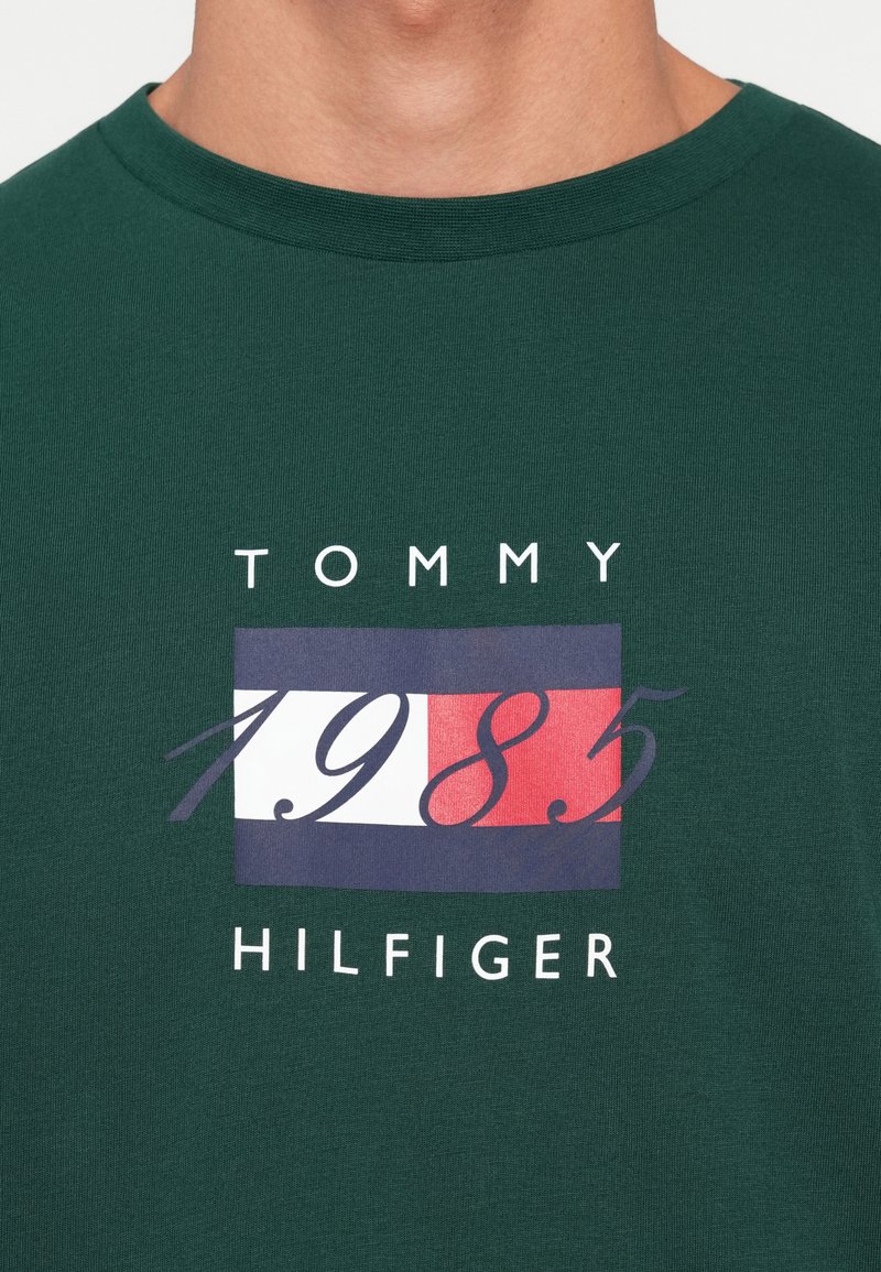 Camiseta de algodón verde con un diseño gráfico que presenta el texto "TOMMY" y "HILFIGER" junto con el año "1985" en rojo y azul sobre un fondo blanco.