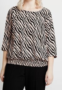 Donna che indossa una blusa larga con motivo a zebra, a tre quarti di manica, e pantaloni neri su uno sfondo neutro.
