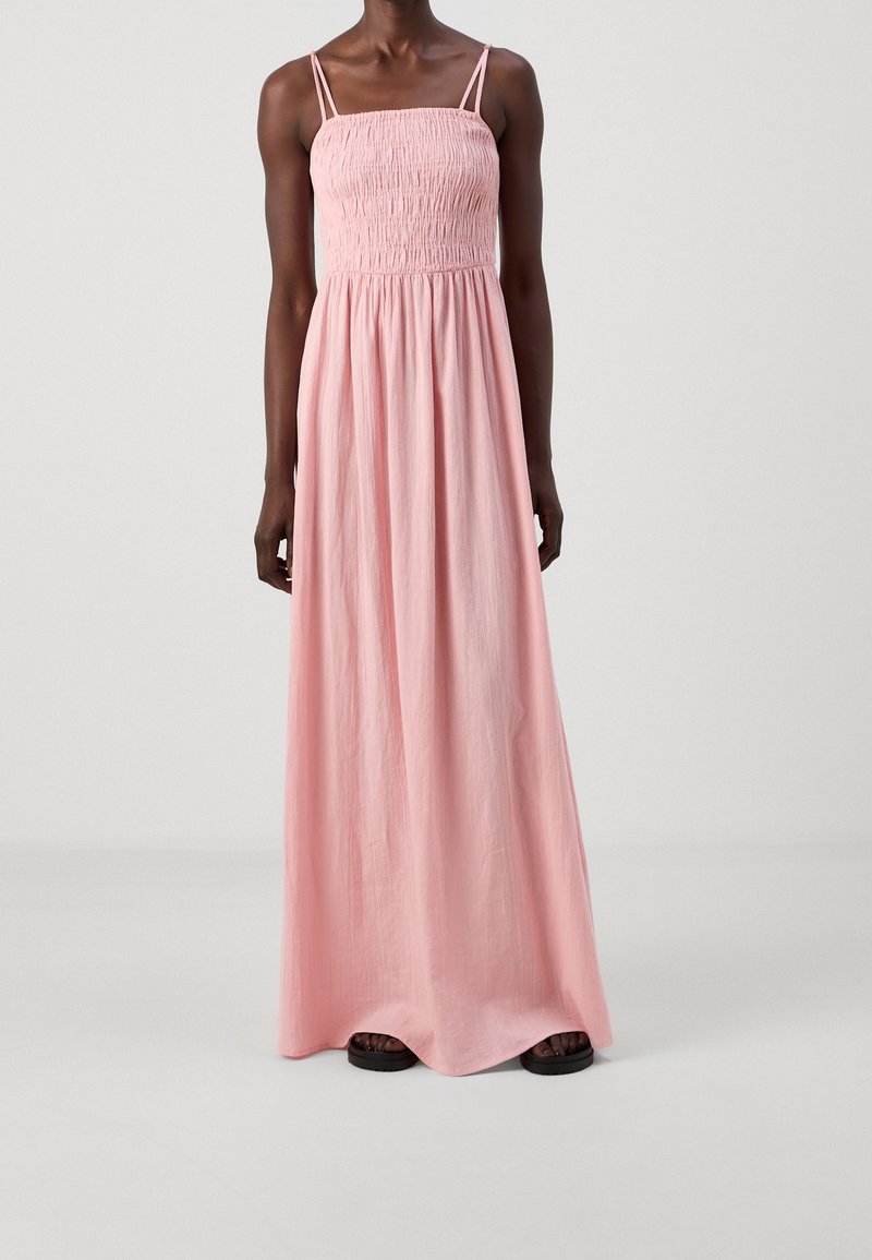 Femme portant une robe longue rose clair sans manches, avec un corsage smocké et des fines bretelles doubles, debout devant un fond uni.