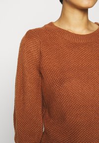 Gebreide trui in roestig oranje met een ronde halslijn, een gewoven patroon en lange mouwen. Het materiaal lijkt zacht en iets losvallend.