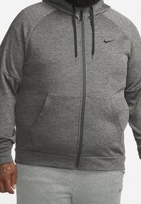 Homme portant un sweat à capuche gris zippé avec des cordons noirs et un pantalon de jogging gris assorti sur fond blanc uni.
