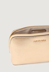 Pungă din piele aurie cu o textură netedă, închidere cu fermoar și branding embosat "MICHAEL KORS" pe față. Formă compactă și rectangulară.