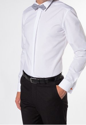 Uomo con camicia bianca, papillon grigio, cintura nera e pantaloni neri, una mano in tasca.
