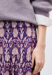Χέρι ακουμπισμένο δίπλα σε φούστα με μοτίβο σε μωβ, κόκκινο και μπεζ, σε συνδυασμό με πλεκτό πουλόβερ λεβάντας και λευκό μανίκι πουκάμισου.