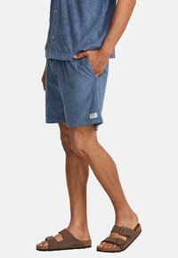 Light blue terry cloth shorts with a raised palm pattern, featuring side pockets. Paired with brown two-strap sandals.