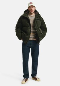 Puffjacke aus dunkelgrünem Cord, mit Kapuze und Druckknopfverschlüssen. Getragen über einem beigen Pullover mit multicoloren Streifen, dunklen Jeans und braunen Sneakers.