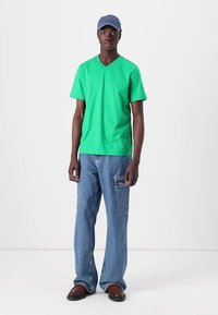 T-shirt vert à col en V, jeans en denim bleu décontractés avec poches latérales, et une casquette bleue. Le mannequin se tient sur un fond blanc. Des chaussures marron complètent le look.