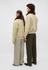 Beige textúrájú dzsekik barna prém gallérral, kockás és csíkos nadrágokkal párosítva. Két alak háttal áll egy semleges háttérben.