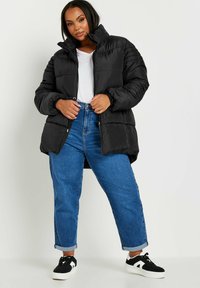 Svart puffjacka med hög krage, med quiltade sektioner och dragkedja i fronten. Kombinerad med uppkavlade blå jeans och svarta sneakers.