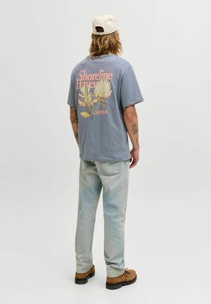 Persona con cappellino beige, maglietta grafica blu con scritta "Shoreline Haven" e motivo floreale, jeans chiari e stivali marroni, vista di spalle.