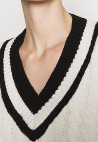 Bílý pletený svetr s hlubokým výstřihem do V, zdobený kontrastním černým žebrovaným lemem. Na hlavní látce je patrný vzor copánkového pletení.