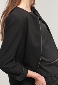 Giacca nera corta con scollo rotondo e cerniera argentata, caratterizzata da una texture liscia e una vestibilità sartoriale, abbinata a una maglia nera sottostante.