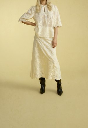 Kobieta w kremowej koronkowej bluzce z rękawami do łokci i pasującej koronkowej spódnicy midi, ubrana w czarne botki na szpic, stojąca na beżowym tle.