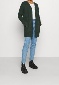 Zelený pletený kardigan s dlouhými rukávy a otevřeným předním dílem, zkombinovaný se světlými modrými džíny a černými šněrovacími kotníkovými botami.