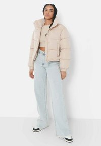 Beige pufferjack met hoge kraag, gestikt ontwerp en zilveren hardware, gecombineerd met lichtblauwe flare jeans en zwart-witte sneakers.