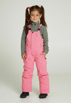 Meisje in roze sneeuwoverall met verstelbare bandjes, voorzakken en een midden-grijze ritsjack. Zwarte schoenen maken de outfit compleet.