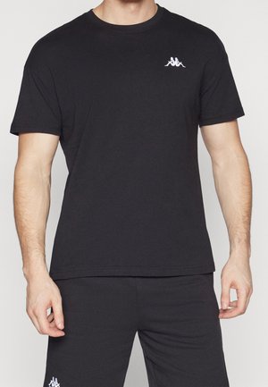 Hombre con una camiseta negra lisa de manga corta y pantalones negros con un pequeño logo blanco en la parte superior izquierda del pecho y el muslo izquierdo.
