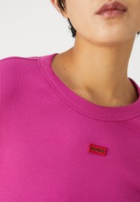 Růžový žebrovaný top s krátkým rukávem a kulatým výstřihem, na hrudi s malou červenou nášivkou "HUGO". Hladká textura a přiléhavý střih.