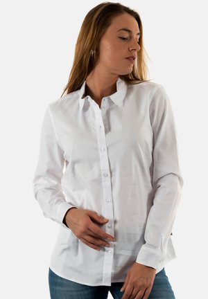 Vrouw in een witte blouse met knopen en een spijkerbroek, die naar beneden en naar links kijkt, terwijl ze de knopen aan de voorkant van de blouse aanraakt.